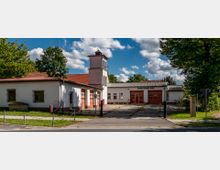 Weißes Feuerwehrgebäude mit roten Toren und einem markanten Turm, umgeben von grünen Bäumen und einem gepflasterten Vorplatz. Im Hintergrund zeigt der Himmel wenige Wolken.