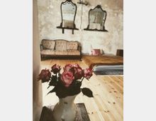 Ein Zimmer mit Holzboden und einer Wand mit abblätterndem Putz, ausgestattet mit einem antiken Sofa, zwei Wandspiegeln mit Holzrahmen und einem niedrigen Bett mit brauner Decke. Im Vordergrund steht eine Vase mit rosa-weißen Rosen auf einem Holztisch.