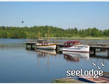 Zwei Boote liegen an einem hölzernen Steg auf einem ruhigen See, umgeben von dichtem grünen Wald und Schilf. Im Vordergrund ist das Logo "seeLodge – Restaurant, Hotel, Seebrücke" eingeblendet.