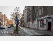 Straßenansicht in einer Stadt mit mehrstöckigen Wohngebäuden aus unterschiedlichen Fassadenstilen. Links führt eine Straße mit Autos, einem Busfahrstreifen und einem Baumbestand entlang, rechts ein schmaler Gehweg mit Geschäften und Straßenschildern, darunter "Tegeler Weg" und "Bonhoeffenufer".