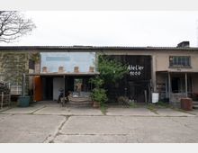 Außenansicht eines einfachen Gebäudekomplexes mit Atelier-Charakter, bestehend aus verputzten und Holzelementen. Ein großer Holztorbereich trägt die Aufschrift „Atelier 1000“, daneben befinden sich ein kleiner Baum, Sitzmöbel, Fahrräder und verschiedene Gegenstände im Außenbereich.