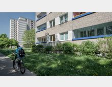 Eine Wohnanlage mit flachen Balkonen und grauer Fassadenoptik, akzentuiert durch blaue Umrandungen. Im Vordergrund führt ein Weg vorbei, auf dem eine Person mit Fahrrad fährt, umgeben von einer Wiese mit Blumen und Bäumen; im Hintergrund steht ein mehrstöckiges Wohnhochhaus.