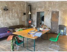 Ein rustikaler Raum mit unverputzten Wänden und Holzboden. In der Mitte steht ein Holztisch mit verschiedenen Gegenständen, umgeben von grünen Stühlen, im Hintergrund ein kleiner Kaminofen sowie eine Couch und eine Holztür.