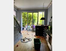 Offener Wohn‑ und Küchenbereich mit Holzfußboden, Treppe links und Küchenzeile rechts an einer weißen Ziegelwand. Große Schiebetür führt auf eine kleine Terrasse mit Gartenblick; im Raum liegen Spielzeug und Bausteine auf dem Boden.