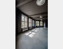 Ein leerer Raum mit modernen, dunklen Wänden, einer grau-schimmernden Betonbodenfläche und großen, rechteckigen Fenstern, die Tageslicht und Schatten auf den Boden werfen. An den Wänden sind Heizkörper unter den Fenstern angebracht, und mehrere Industrieleuchten hängen von der Decke.