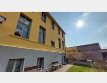 Gelbes Wohngebäude mit großen Fenstern, dessen unterer Bereich grau verputzt ist. Im Vordergrund befindet sich ein gepflasterter Weg, eine Rasenfläche und eine Sitzbank, im Hintergrund steht ein gemischtes Gebäude aus Holz und Backstein unter blauem Himmel mit Sonne.