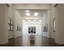 Minimalistisch gestalteter Ausstellungsraum mit hellen Wänden und glänzendem Boden. An den Seitenwänden hängen gerahmte Schwarz-Weiß-Fotografien, während in der Mitte ein schmaler Holztisch steht.