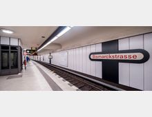 U-Bahn-Station „Bismarckstraße“ mit weiß verkleideten Wänden, schwarzen Kreuzmotiven und roten Schriftzügen. Die Gleise verlaufen parallel zum Bahnsteig, der mit Wegemarkierungen versehen ist; eine Anzeigetafel und eine Wanduhr befinden sich im Hintergrund.