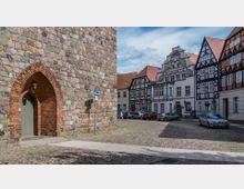 Historischer Platz mit Kopfsteinpflaster, umgeben von Fachwerkhäusern und barockem Giebelhaus; links ein großes Gebäude aus Naturstein mit einem gotischen Torbogen aus roten Ziegeln. Im Vordergrund parkende Autos.