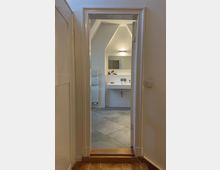 Blick durch eine Tür in ein modernes Badezimmer mit grauen Fliesen, einem weißen Waschbecken mit Wandarmatur, einem beleuchteten Spiegel und einem Handtuchheizkörper an der linken Wand. Die Wände sind weiß, und die Schräge der Decke ist sichtbar.