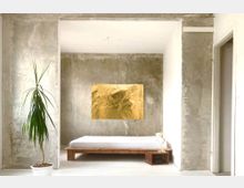 Minimalistisches Schlafzimmer mit rohen Sichtbetonwänden, einem schlichten Holzbett mit weißem Bettzeug und einem kleinen Holztisch mit Lampe. Links im Raum steht eine grüne Zimmerpflanze in einem weißen Topf, an der Wand hängt ein goldenes Gemälde.