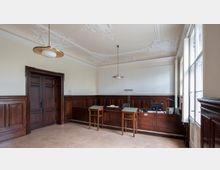 Ein Raum mit holzvertäfelten Wänden, einer Holz-Doppeltür und einer mit Holz verkleideten Theke. Die Decke ist mit aufwendigen Stuckmustern verziert, und an der rechten Seite befinden sich hohe Fenster, die viel Tageslicht hereinlassen.