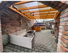 Überdachter Hofbereich mit Ziegelfassaden, einer Sitzgruppe aus grauem Flechtmaterial und einem Holztisch. Im Hintergrund ein Holzheizfass sowie gestapeltes Brennholz, ergänzt durch eine klare Kunststoffdachabdeckung mit Holzstreben.