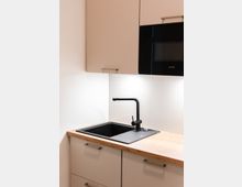 Minimalistische Küchenecke mit einer hellen Arbeitsplatte aus Holz, einem schwarzen Spülbecken mit passender Armatur, hellen Unterschränken mit Metallgriffen und einem Hängeschrank, der teils eine eingebaute schwarze Mikrowelle enthält.