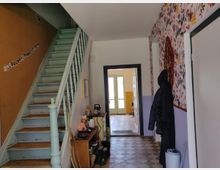 Flur in einem Wohnhaus mit einer hellblauen Holztreppe auf der linken Seite. Rechterhand eine Garderobe vor einer Blumenmuster-Tapete, sowie eine Türöffnung, die in einen weiteren Bereich mit Fliesenboden und doppeltürigen Fenstern führt. Entlang der Treppe hängen kleine Fotos an der Wand.
