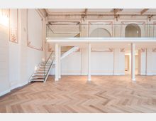 Heller, hoher Innenraum mit Fischgrät-Parkettboden, weißen Wänden mit dekorativen Stuck- und Ornamentrahmen sowie einer eingezogenen Galerieebene mit Glasgeländer. Links führt eine moderne Treppe mit Glasbrüstung zur oberen Ebene; im Hintergrund sind bogenförmige Durchgänge und Stützen zu sehen.