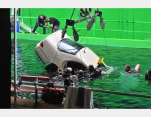 Szene einer Filmproduktion in einem Wasserbecken mit grünem Hintergrund (Greenscreen); ein teilweise ins Wasser eingetauchtes Auto ist umgeben von mehreren Tauchern und technischem Equipment. Im Vordergrund sind Ausrüstungen und Handtücher zu sehen.