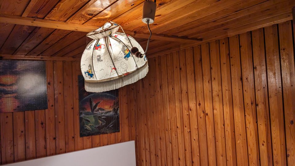 Innenansicht eines kleinen Zimmers mit holzvertäfelten Wänden und Holzdecke. An der Decke hängt eine gemusterte Pendelleuchte, an der Wand sind zwei Bilder und am unteren Bildrand ist ein weißes Bettkopfteil zu sehen.