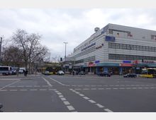 Straßenecke in einer Stadt mit einem großen, mehrstöckigen Gebäude im Hintergrund, an dessen Fassade Schilder von Geschäften und Fahnen angebracht sind. Im Vordergrund befindet sich eine belebte Kreuzung mit mehreren Autos, gelben Doppeldeckerbussen und Fußgängern; kahle Bäume säumen den Straßenrand.