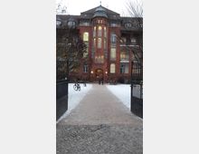 Rotes Backsteingebäude mit symmetrischer Fassade, mittigem Turmerker und erleuchteten Fenstern, gelegen hinter einem schneebedeckten Gehweg, der durch ein offenes Eisentor führt. Im Vordergrund sind Fahrradständer und einzelne Personen sichtbar.