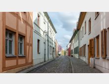 Schmale, gepflasterte Gasse mit historischen Häusern auf beiden Seiten; links ein Fachwerkgebäude, rechts Gebäude mit hölzernen Fensterläden. Am Ende der Straße sind blühende Bäume und blauer Himmel erkennbar.