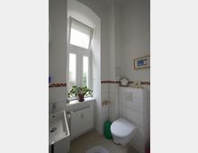 Kleines Badezimmer mit weißen Wänden und Fliesen, einer Toilette, einem kleinen Waschbecken und einem großen, halb offenen Fenster mit einer Pflanze auf der Fensterbank. Unter dem Fenster befindet sich ein Heizkörper, und an der Wand hängt ein gerahmtes Bild.