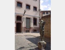 Der Innenhof eines historischen Backsteingebäudes mit weißen Wänden und vergitterten Fenstern. Im Vordergrund steht eine skulpturale Bronzefigur auf einem kleinen Sockel, umgeben von Pflastersteinen.