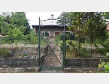 Grünes Metall-Gartentor mit Maschendraht und Hausnummer „15“ in einem Zaun, dahinter ein schmaler Weg durch einen dicht bewachsenen Vorgarten zu einem kleinen, eingeschossigen Haus. Umgeben von Bäumen und Sträuchern, mit Briefkasten am Zaun und niedriger Steinmauer entlang der Grundstücksgrenze.