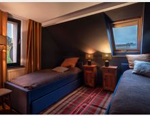 Schlafzimmer mit zwei Einzelbetten in dunkelblauer Polsterung, flankiert von zwei Nachttischen aus Holz mit jeweils einer Tischlampe. Links ein Fenster mit Vorhang, rechts ein Dachfenster, und ein gemusterter Teppich bedeckt den Boden.