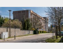 Wohnstraße in einer Vorstadt mit mehrstöckigem Wohngebäude aus Beton im Hintergrund, umgeben von Bäumen und Sträuchern. Auf der rechten Seite stehen Straßenschilder, geparkte Autos und Gehwege.