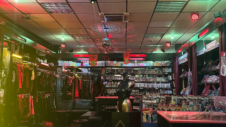 Ein Verkaufsraum mit niedriger Rasterdecke, bunter Beleuchtung und Schaufensterpuppen. Regale an den Wänden präsentieren Kleidung und Accessoires, während Glasvitrinen kleine Artikel ausstellen.