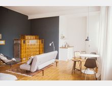 Wohnraum mit gemütlichem, modernem Design: Ein helles Zimmer mit Holzboden, einer hellgrauen Couch, einem zierenden Teppich und einem großen, goldfarbenen Kachelofen an einer dunkelblauen Wand. Im Hintergrund befinden sich eine kleine Küchenzeile und ein runder Esstisch mit verschiedenen Stühlen.