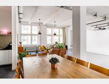 Heller, offener Raum in einem modernen Loft-Stil mit weiß gestrichenen Wänden, großen Fenstern und sichtbaren Deckenträgern. Im Vordergrund ein großer Esstisch aus Holz mit Stühlen, dahinter eine Sitzecke mit Holzmöbeln, Pflanzen und einer kleinen Teeküche auf der linken Seite.