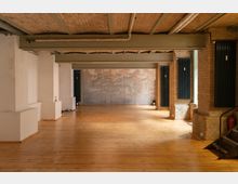 Ein geräumiger, rechteckiger Innenraum mit Holzboden und einer Decke aus freiliegenden Backsteinen. Die Wände bestehen aus einer Kombination von weiß verputztem Mauerwerk und gelben Ziegelsteinen, mit großflächigen Heizkörpern an den Seiten. Im Hintergrund ist eine Wand mit einem gedruckten Waldmotiv zu sehen.