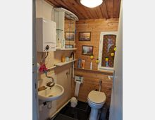 Kleines Badezimmer mit Holzvertäfelung und schräger Holzdecke, ausgestattet mit Waschbecken, wandhängendem Boiler, Spiegel-Schrank und WC. An der Rückwand befindet sich ein kleines Fenster mit gemustertem Glas sowie Regale mit Toilettenartikeln und gerahmten Bildern.