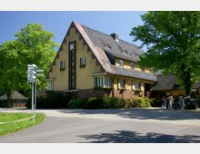 Großes Gebäude mit gelber Fassade und steilem Satteldach aus dunklen Schindeln, ergänzt durch Backsteinelemente und eine vertikale Uhr im Giebel. Umgeben von Bäumen und Grünflächen, mit einem asphaltierten Weg und einem Wegweiser im Vordergrund.