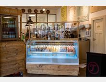 Innenraum einer Eisdiele mit einer beleuchteten Kühlvitrine voller verschiedener Eissorten auf Stielen. Die Einrichtung besteht aus Holzelementen, dahinter befinden sich eine Kaffeemaschine und Wanddekorationen mit Speisekarten.