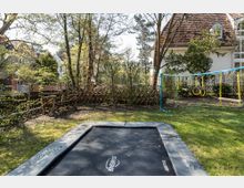 Garten mit einem Bodentrampolin im Vordergrund, umgeben von Rasen und Bäumen. Im Hintergrund stehen ein Klettergerüst mit Schaukeln sowie ein Haus mit roten Dachziegeln und mehreren Fenstern.