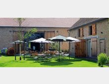 Ein Außenbereich mit grüner Rasenfläche zwischen zwei Gebäuden aus Backstein mit Holztüren und -fenstern, eingerichtet mit mehreren Tischen und Stühlen unter großen Sonnenschirmen. Im Hintergrund ist ein offenes Scheunentor zu sehen.