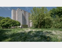 Hochhaus mit schlichter Fassade aus horizontalen Streifen, umgeben von einer Grünfläche mit blühendem Wiesengrund und mehreren großen Bäumen; im Hintergrund dichter Baumbewuchs.