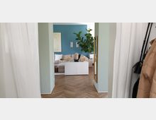 Wohnraum mit pastellgrünen Wänden und einer akzentuierten blauen Wand im Hintergrund, darauf minimalistische Kunstwerke. Im Vordergrund ein heller Holzboden in Fischgrätenmuster sowie ein weißes Sideboard mit Deko-Objekten, darunter eine Büste und zwei Korblampen. Im hinteren Bereich ein beigefarbenes Sofa, Pflanzen und weitere moderne Einrichtungselemente.