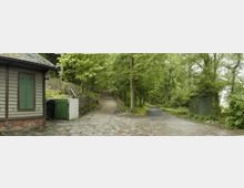 Kopfsteinpflasterweg mit einer Weggabelung in einem bewaldeten Gebiet. Links ist ein kleines Gebäude mit grün gestrichenen Fensterläden und rechts ein grüner Stromkasten, umgeben von dichten Bäumen und grüner Vegetation.