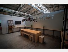 Moderner, heller Innenraum mit industriellen Deckenstrukturen, einem langen Holztisch mit Hockern in der Mitte und Regalen sowie Pflanzen an den Wänden. An der Rückwand leuchtet ein Schild mit der Aufschrift "FEINKOST", begleitet von einer Hängeleuchte aus mehreren kleinen Lampen.