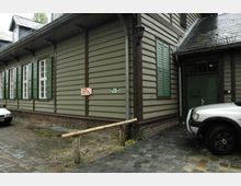 Außenansicht eines Gebäudes in Holzbauweise mit grün gestrichenen Fensterläden und einer Eingangstür. Im Vordergrund Pflastersteine, ein geparktes Auto und Hinweisschilder mit der Aufschrift „Zu den Toiletten“ und „Eingang zum Bierkeller“.