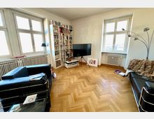 Wohnzimmer mit Parkettboden im Fischgrätmuster, weißen Wänden und großen Fenstern. Der Raum enthält schwarze Ledersofas, ein weißes Bücherregal, einen Flachbildfernseher auf einem Stativ sowie Heizkörper unter den Fenstern.