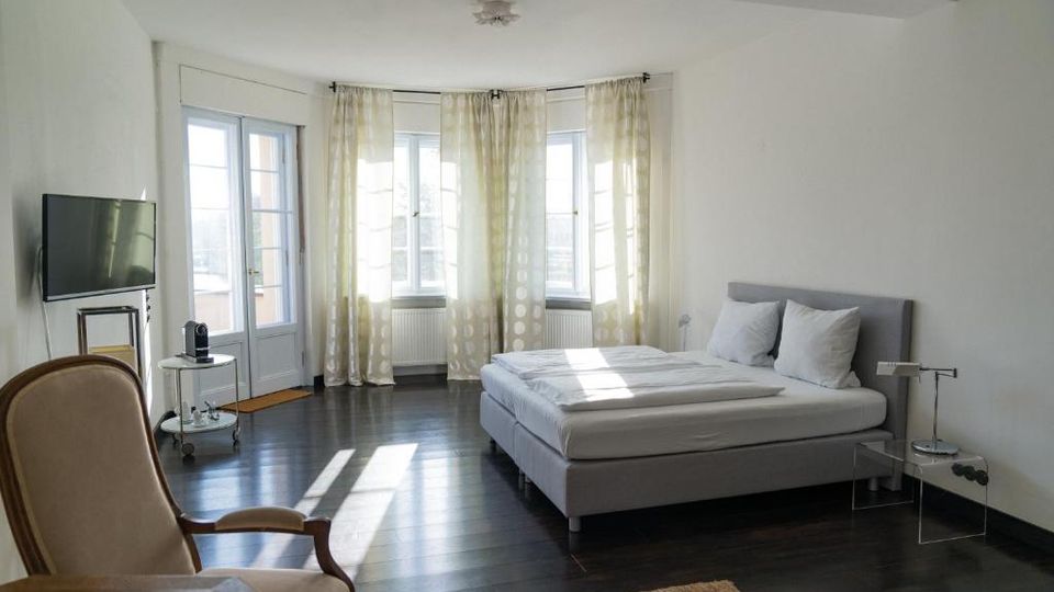 Helles, minimalistisches Schlafzimmer mit Doppelbett, dunklem Holzboden und großen Fenstern mit hellen Vorhängen in einer gebogenen Fensterfront. Links stehen ein Fernseher und ein kleiner Beistelltisch, daneben eine Glastür mit Fensterflügeln.