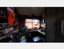 Ein kleiner Arbeitsraum mit Regalwänden voller Gegenstände wie Werkzeuge, Kartons und Geräte. Ein Fenster mit beigefarbenen Vorhängen beleuchtet den Raum, daneben ein Schreibtisch mit weiteren Utensilien und einer kleinen Kaffeemaschine.