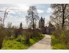 Ein historisches Schloss mit spitzen Türmen und roten Fassadenelementen, umgeben von Bäumen, Sträuchern und Wiesen. Ein schmaler Wanderweg führt durch die Grünfläche zum Gebäude, während der Himmel leicht bewölkt ist.
