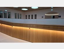 Innenansicht eines modernen Foyers oder Flurs mit geschwungener Brüstung aus Holz und indirekter Beleuchtung. Helle Wände mit mehreren Fenstern, ein großer Rundbogen-Durchgang sowie runde Deckenleuchten unter einer perforierten Decke prägen den Raum.