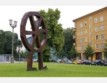 Eine große, abstrakte Skulptur aus rostfarbenem Metall in Form eines Rads steht auf einer Wiese, umgeben von Bäumen. Im Hintergrund sind ein gelbes Wohngebäude mit Balkonen, parkende Autos und eine Straßenlaterne zu sehen.
